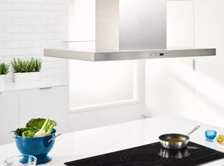 Cucina bianca moderna con cappa sopra il piano induzione nero, pentola sul fornello e ciotola azzurra con verdure.
